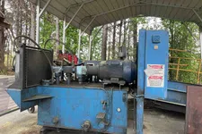 Thumbnail for Industrial blue hydraulic machine with electric motor under a metal canopy outdoors surrounded by trees.