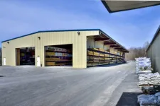 Thumbnail for Large metal warehouse with open storage racks holding lumber and piles of bags outside on paved ground under blue sky