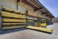 Thumbnail for Yellow forklift carrying long wooden planks outside a lumber store with stacks of wood on metal racks under roof.