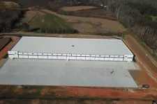 Thumbnail for Aerial view of a large new industrial warehouse with a white roof surrounded by cleared land and forest.