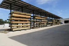 Thumbnail for Outdoor lumber yard with stacked wooden planks under a metal canopy on a sunny day