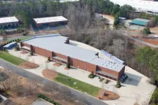 Thumbnail for Aerial view of a large brick commercial building with a metal roof surrounded by trees and parking areas.