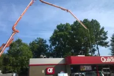 Thumbnail for Concrete pump truck extending long arm over QuikTrip store with trees and blue sky in background