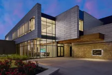 Thumbnail for Modern commercial building with glass walls, stone facade, and illuminated interior at twilight under a purple sky.