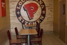 Thumbnail for Interior of a Popeyes Louisiana Kitchen with wooden tables, chairs, and large logo mural on wood wall.