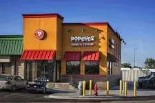 Thumbnail for Exterior view of a Popeyes Louisiana Kitchen fast-food restaurant with bright orange and red facade under clear blue sky.
