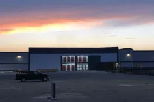 Thumbnail for Warehouse building with bright entrance lights and a black pickup truck in a large empty parking lot at sunset.