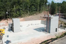 Thumbnail for Construction site with concrete foundation and walls in early stages surrounded by trees and construction equipment.