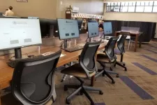 Thumbnail for Row of modern computer stations with black ergonomic chairs in a public library setting with bookshelves and study area.