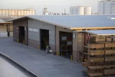 Thumbnail for Outdoor lumber storage racks beside a warehouse building with industrial tanks in the background under a clear sky.