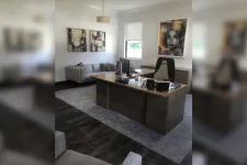 Thumbnail for Modern office with wooden desk, black chair, couch, abstract art, and a large window with white walls.