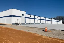 Thumbnail for Large white industrial warehouse with a blue stripe under clear sunny sky, surrounded by dirt and gravel.