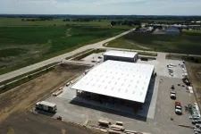 Thumbnail for Aerial view of a large warehouse with white roof surrounded by farmland and vehicles on a sunny day
