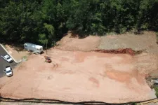Thumbnail for Aerial view of cleared construction site with dirt, parked vehicles, and surrounding trees in daylight.