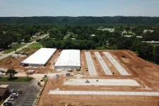 Thumbnail for Aerial view of a construction site with two large warehouse buildings and concrete slabs surrounded by dirt and greenery.