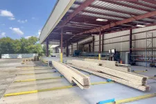 Thumbnail for Stacked lumber under a metal roofed warehouse with clear sky and trees in the background