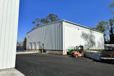 Thumbnail for White industrial warehouse with forklifts and stacked pallets on asphalt under clear blue sky.
