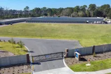 Thumbnail for Aerial view of a gated self-storage facility with multiple storage units and large green lawn under clear sky.