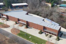 Thumbnail for Aerial view of a large brick industrial building with a metal roof surrounded by parking areas and trees.