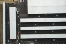 Thumbnail for Aerial view of multiple long white-roofed warehouse buildings with black asphalt roads and parking areas.