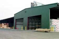 Thumbnail for Large green warehouse storing various wood planks and construction materials with open bays.