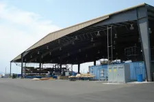 Thumbnail for Large open warehouse structure with metal roof, containers, and industrial materials on concrete ground under clear sky