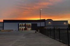 Thumbnail for Large modern building at sunset with illuminated windows and empty parking lot under colorful sky.