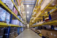 Thumbnail for Wide aisle in warehouse lined with tall yellow shelves stocked with boxes and packaged goods.