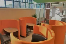Thumbnail for Modern office lounge area with curved orange seating pods, small wooden tables, and black chairs under natural light.
