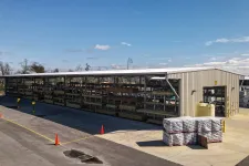 Thumbnail for Outdoor lumber storage rack under metal shelter with pallets and an industrial warehouse in a sunny setting.