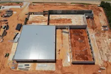 Thumbnail for Aerial view of a construction site with a large metal-roofed building and adjacent concrete foundation walls on red soil.