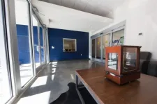 Thumbnail for Modern office waiting area with large glass doors, blue accent wall, gray couch, and wooden table with case.