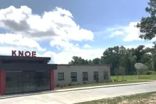 Thumbnail for Exterior view of a small building with KNOE sign on the roof under a blue sky with clouds and surrounding greenery.