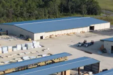 Thumbnail for Aerial view of a lumberyard with stacked wood, large storage buildings, and trucks under blue roofs.