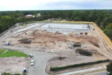 Thumbnail for Aerial view of a large construction site with earthworks, concrete slabs, equipment, and surrounding trees under a cloudy sky