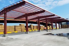 Thumbnail for Red steel frame structure under construction in an outdoor lumber yard with stacked wooden planks and clear sky.