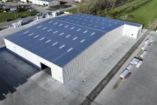 Thumbnail for A large industrial warehouse with a blue roof and surrounding concrete yard in an open area.