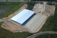 Thumbnail for Aerial view of a large white industrial warehouse surrounded by cleared land and greenery with adjacent roads.