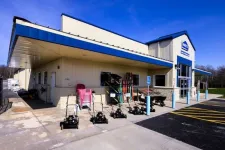 Thumbnail for Exterior view of a hardware store with lawnmowers and garden tools displayed outside under sunny blue sky.