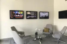 Thumbnail for Modern waiting area with four gray chairs around a glass coffee table and three wall-mounted TVs.