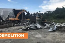 Thumbnail for Two excavators demolishing a large metal building with debris scattered around on a construction site.