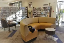 Thumbnail for Modern library interior with curved beige sofa, bookshelves, armchair, and large windows letting in natural light