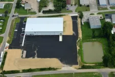 Thumbnail for Aerial view of industrial warehouse with large parking lot and adjacent pond surrounded by greenery and roads.