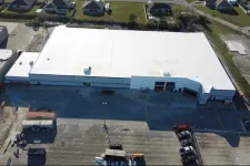 Thumbnail for Aerial view of a large white-roofed commercial warehouse building with surrounding parking and nearby houses.
