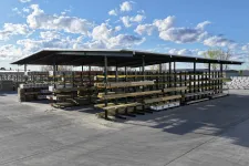 Thumbnail for Outdoor lumber storage with organized metal racks under a metal roof on a sunny day with scattered clouds.