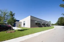 Thumbnail for Modern single-story commercial building with gray walls, green lawn, parked cars, and clear blue sky.