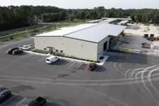 Thumbnail for A large industrial warehouse with several parked vehicles and tire marks in a spacious asphalt lot surrounded by trees.