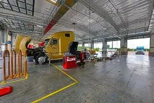 Thumbnail for Large yellow truck undergoing maintenance inside a spacious, well-lit truck service garage with equipment and open bays.