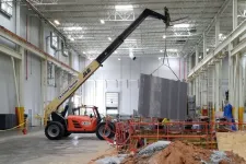 Thumbnail for Orange JLG telehandler lifting a large metal panel inside a spacious industrial warehouse under construction.