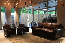 Thumbnail for Cozy living room with brown leather sofa, two armchairs, round black table, rug, and large glass doors to outside.